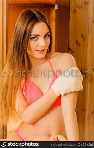 Young woman in wood finnish spa sauna massaging skin with exfoliating glove. Girl in bikini relaxing. Skincare concept.. Woman in sauna with exfoliating glove. Skincare.