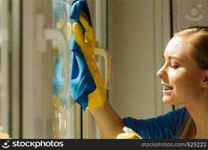Young woman in yellow gloves cleaning window with blue rag and spray detergent. Spring cleanup, housework concept. Girl cleaning window at home using detergent rag