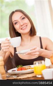 Young woman is drinking a hot cup of coffee while having a breakfast at home