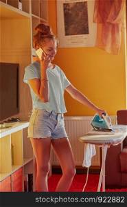 Young woman is ironing her clothes and talking on a smartphone simultaneously, standing by a ironing board in a room at home. Routine housekeeping task at home. Candid people, real moments, authentic situations
