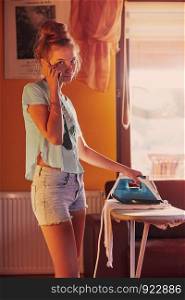 Young woman is ironing her clothes and talking on a smartphone simultaneously, standing by a ironing board in a room at home. Routine housekeeping task at home. Candid people, real moments, authentic situations