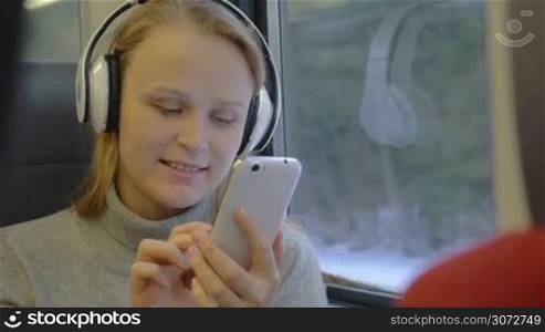 Young woman is listening to the music in headphones and writing messages in her smartphone while traveling by train.