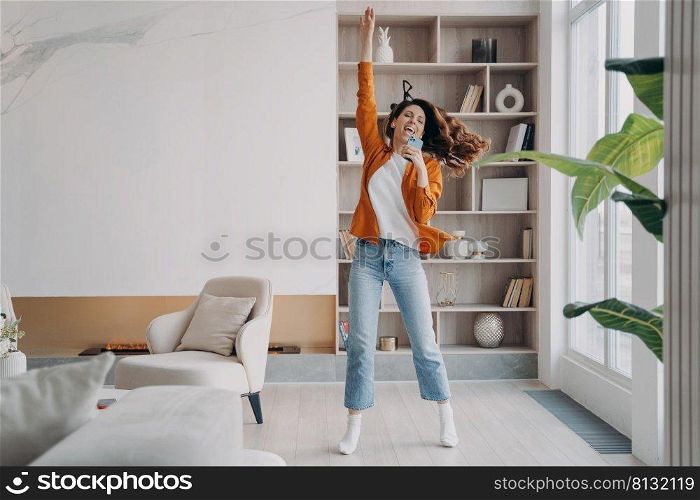 Young woman is singing, dancing and jumping. Carefree girl with smartphone having fun crazy and screaming at home. Favourite song and relaxation. Concept of emotional expressions and rebellion.. Young woman is singing, dancing and jumping. Girl having fun crazy and screaming at home.