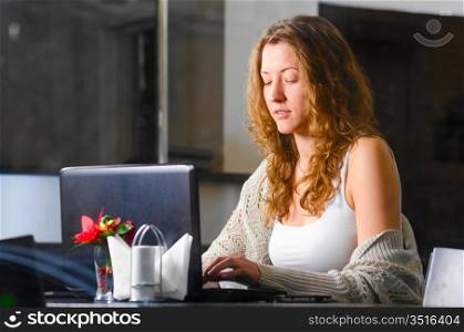 young woman is sitting at cafe and working on laptop. View through the window glass from the outside.
