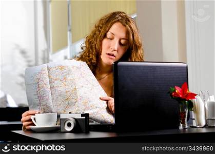 young woman is sitting at cafe with laptop and looking at road map