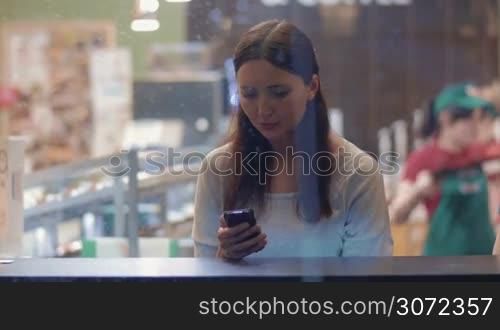 Young woman is sitting in cafe and writing messages to friends in smartphone. Shot is made through the cafe show window.