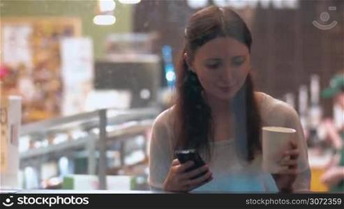 Young woman is sitting in coffee shop, drinking coffee and texting to friends using smartphone. Shot is made through the show window.