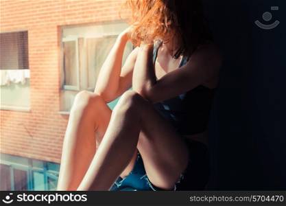 Young woman is sitting in the sunlight on a window sill and looking out