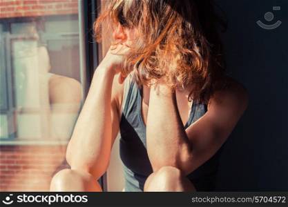 Young woman is sitting in the sunlight on a window sill and looking out