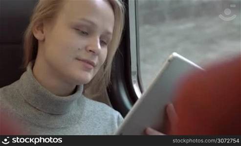 Young woman is sitting in train carriage by window. She&acute;s typing something in her tablet PC and smiling.