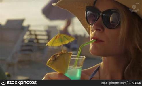 Young woman is sitting on the beach in sun hat and sunglasses and drinking cocktail through the straw.