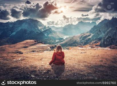 Young woman is sitting on the field against the majestic mountains at sunset in autumn in Dolomites, Italy. Landscape with girl, cloudy sky, orange grass, high rocks with forest in fall. Italian alps