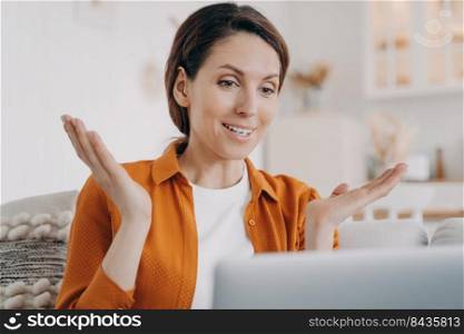 Young woman is speaking online and gesticulating. European girl has online conference at home. Happy lady in orange shirt with laptop is talking at internet meeting. Working while quarantine.. Happy young woman is speaking online and gesticulating. European girl has online conference at home.