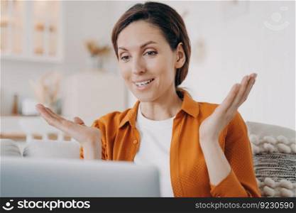 Young woman is speaking online and gesticulating. European girl has online conference at home. Happy lady in orange shirt with laptop is talking at internet meeting. Working while quarantine.. Happy young woman is speaking online and gesticulating. European girl has online conference at home.