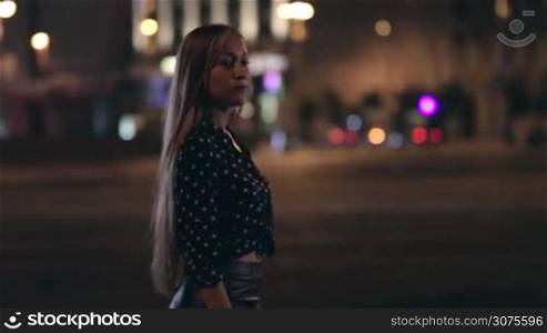 Young woman is strolling alone on city streets at night. On background cityscape and city lights