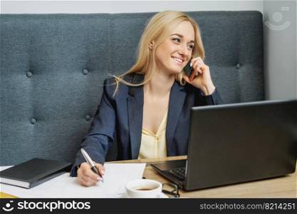 Young woman is talking by cell phone sitting at the table in home office.. Young woman is talking by cell phone in office.