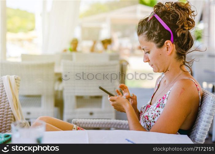 Young woman is using mobile smart phone to send or read message or to make a call on social network during a break at the cafe or restaurant in sunny day outdoors