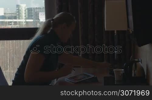 Young woman is working at table using tablet PC. She&acute;s turning the desk lamp on, answer the phone and drinking beverage from the cup.