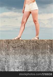 Young woman legs wearing sandals summer shorts standing on stone wall against sky