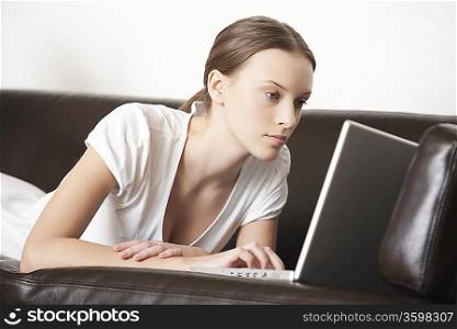Young woman lies on leather sofa working on laptop
