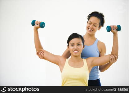 Young woman lifting hand weights while another woman helps position her arms.