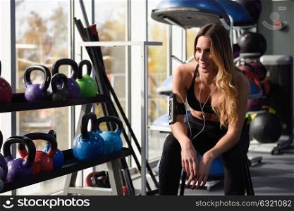 Young woman listening to music with smartphone sitting at gym. Young fitness female using headphones with dumbbells at the background.