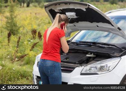Young woman looking at broken car and talking by phone