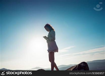 Young woman looking at her phone against sky, while standing on a rock