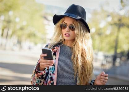 Young woman looking at her smartphone in urban background. Girl wearing jacket, hat, sweater and sunglasses.