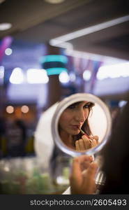 Young woman looking in handheld mirror while applying lipstick