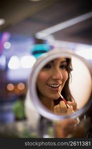 Young woman looking in handheld mirror while applying lipstick