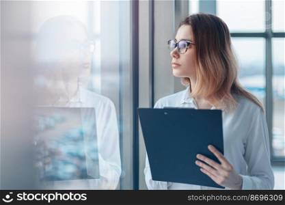 Young woman looking in the panoramic window of a modern office and holding a folder with documents.