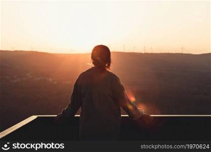 Young woman looking the mountains from a balcony during a super colorful sunset with copy space, freedom and wellness concept