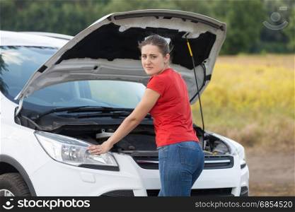 Young woman looking under the hood of broken car at rural road