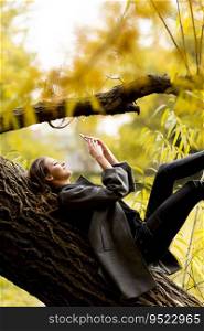Young woman lying on a branch in the autumn park with phone in hand