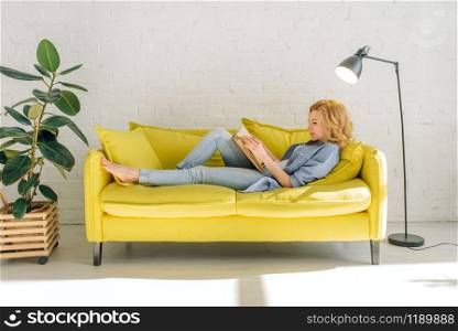 Young woman lying on cozy yellow couch and reading a book, living room in white tones on background. Attractive female person with magazine sitting on sofa at home. Woman lying on cozy yellow couch and reading book