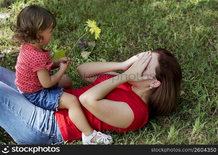 Young woman lying on grass and covering her face with her hands and her daughter sitting on her stomach
