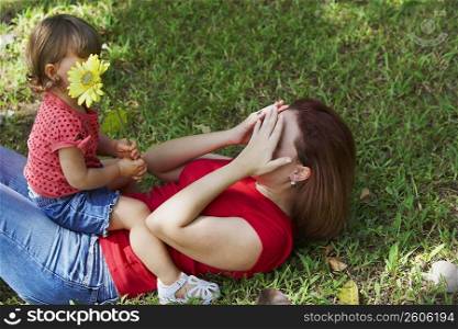 Young woman lying on grass and covering her face with her hands and her daughter sitting on her stomach