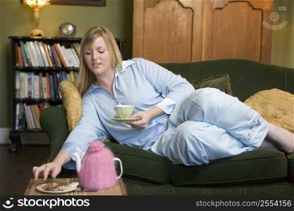 Young woman lying on sofa at home eating cookies and drinking tea