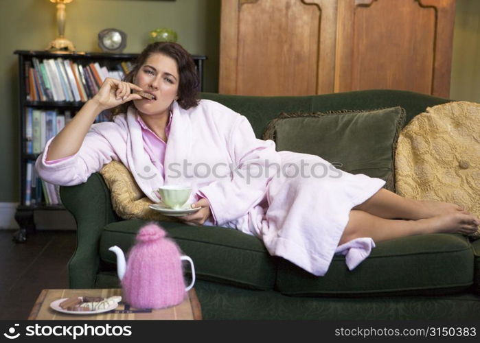 Young woman lying on sofa at home eating cookies and drinking tea