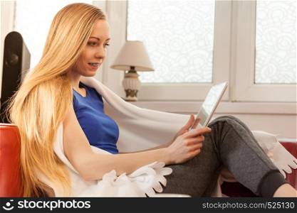 Young woman lying on sofa under warm blanket, relaxing using tablet, reading news and checking social media, having fun during leisure time. Modern relaxation.. Young woman relaxing with tablet