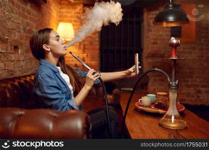 Young woman makes selfie with hookah in bar, mesmerizing cloud. Shisha smoking, traditional bong culture, tobacco aroma for relaxation, rest with hooka. Young woman makes selfie with hookah in bar