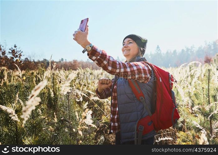 Young woman making video call sending greetings from vacation trip in mountains. Woman with backpack hiking through tall grass along path on meadow. Spending summer vacation close to nature