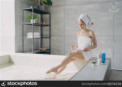 Young woman massaging legs with wooden brush sitting on bathtub wearing white towel. Female wrapped in towels massages hips to prevent cellulite. Body care, spa procedure in bathroom.. Young woman massaging hips to prevent cellulite sitting on bathtub. Body care, spa procedure