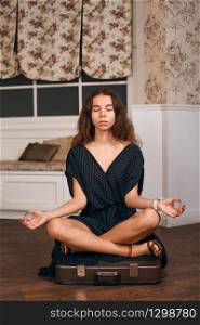 Young woman meditates in a pose of yoga on a suitcase. Retro styled living room on the background.. Young woman sits in a pose of yoga on suitcase.