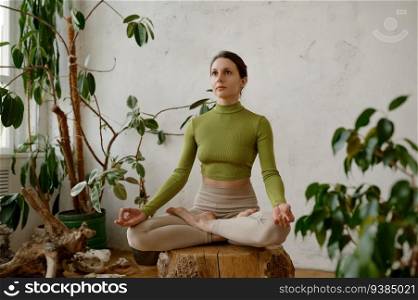 Young woman meditating on wooden stump practicing yoga indoors. Calm mindful female yogi breathing, harmonizing internal and external space, body, mind, soul and thoughts. Young woman meditating on wooden stump practicing yoga indoors