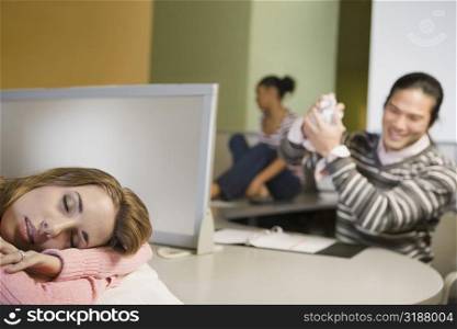 Young woman napping on a desk with a young man smiling in the background