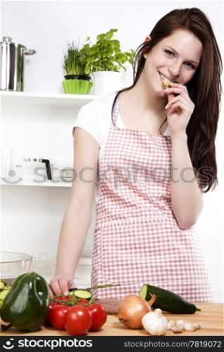 young woman nibbling on a cucumber. happy young woman in a kitchen nibbling on a cucumber while chopping for salad