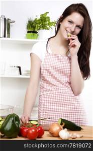 young woman nibbling on a cucumber. happy young woman in a kitchen nibbling on a cucumber while chopping for salad
