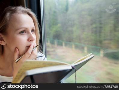 Young woman on a train writing notes in a diary or journal staring thoughtfully out of the window with her pen to her lips as she thinks of what to write